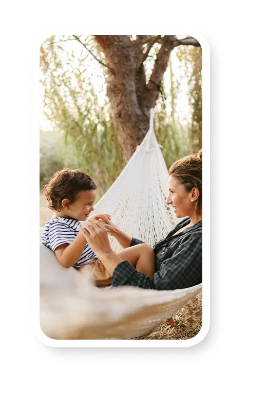 A mother and her child sitting in a hammock outside and smiling at each other.